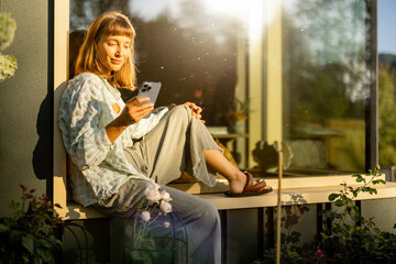 A woman relaxes by a window in a sunlit garden, checking her phone. Surrounded by blooming flowers, she enjoys a moment of solitude and peace, escaping to nature
