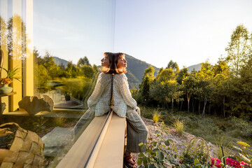 A woman sits quietly by a window, gazing at the surrounding mountains and forest. The serene...