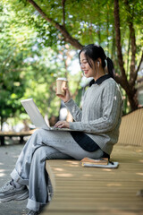 A woman enjoys a hot drink while working remotely outdoors, sits on a park bench with her laptop.