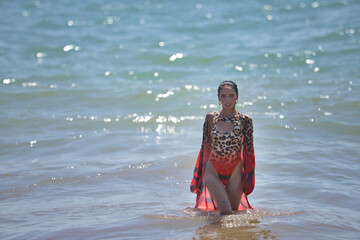 Fototapeta premium Young woman, beautiful, brunette, with a leopard swimsuit and sarong, totally wet, walking out of the water on the shore of the beach with the sea in the background. Concept beauty, femininity.