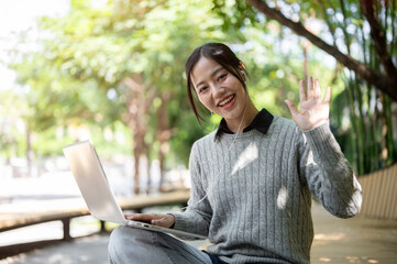 Fototapeta premium A beautiful Asian woman sits on a park bench with her laptop, smiles warmly and waves at the camera.