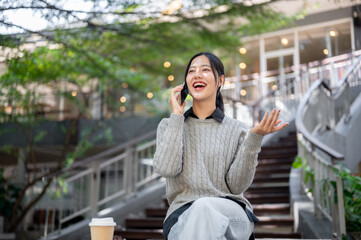 A cheerful, smiling Asian woman enjoys talking on the phone while sitting outdoors in the city.