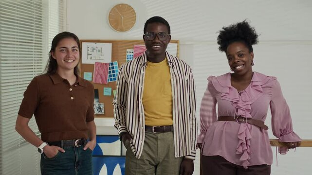 Medium portrait of multiethnic team of three smiling at camera while standing against corkboard with drafts and notes on it, working together in contemporary creative workspace