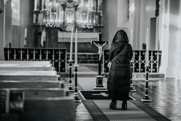 A person in a hooded coat standing before a crucifix in a church, with a solemn ambiance, candles,...