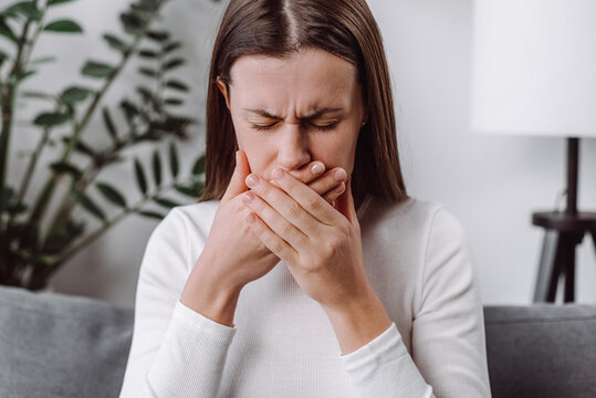 Close up of young woman suffering from nausea at home, caucasian female having morning sickness, ill lady sitting alone on couch in living room, covering mouth