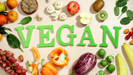 The word VEGAN in large green letters on beige table, surrounded by fresh fruits, vegetables, and a bowl of lentils