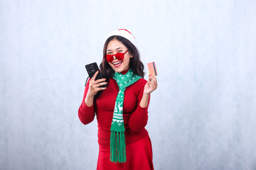 Asian adult girl happy to camera holding gadget screen, tilted credit debit card, wearing glasses, wearing red christmas sweater with Santa hat and scarf, isolated on white background