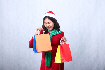 gesture of young asian girl wearing red christmas sweater, santa hat and scarf, cheerful looking at bag contents both hands lifting colorful bags isolated on white background