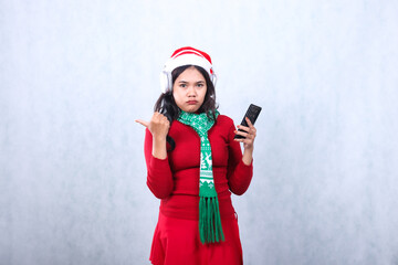 expression of young woman wearing christmas sweater, headphone, santa hat and scarf, pouting to camera hand holding hand phone, cellphone, smart phone and pointing right, isolated on white background