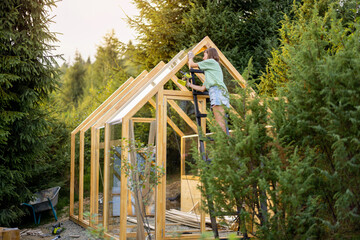Female carpenter using a power drill while working on a wooden greenhouse in her backyard, engaged...