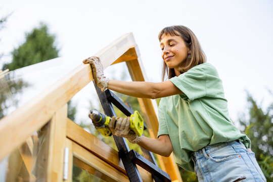 Female carpenter using a power drill while working on a wooden greenhouse in her backyard. She is wearing casual clothes and work gloves, engaged in a DIY project, surrounded by nature