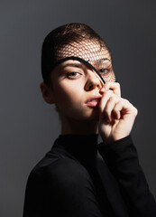 Elegant woman with fishnet veil holding hand near face, dressed in black, showcasing strong expression. Fashion and beauty concept in a minimalistic setting.