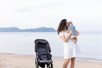 A loving mother kisses her baby while standing on a sandy beach next to a stroller. The calm sea...
