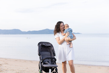A loving mother kisses her baby while standing on a sandy beach next to a stroller. The calm sea...