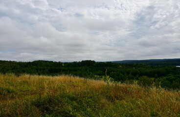 A view of a hill or mountain covered from all sides with grass, reeds, forests, and moors, located next to a vast river or lake, meadows, as well as having a view on nearby villages and towns 