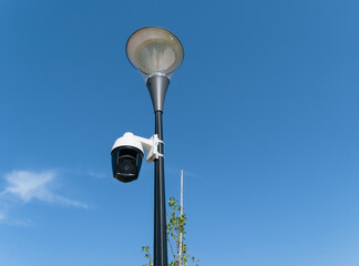 Security video camera mounted on a lamppost outdoors against a blue, partly cloudy sky