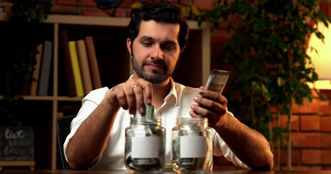 Indian bearded young man sitting at a desk under moody lighting, managing finances by putting money into two glass jars with blank labels for customisation, symbolising saving and financial planning