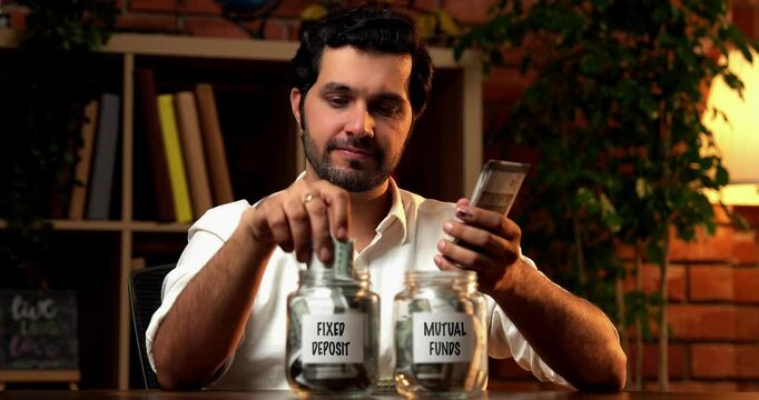 Indian bearded young man managing finances by putting money into a glass jar labeled for fixed deposits and mutual funds, symbolizing savings and investment planning, sitting at home on desk