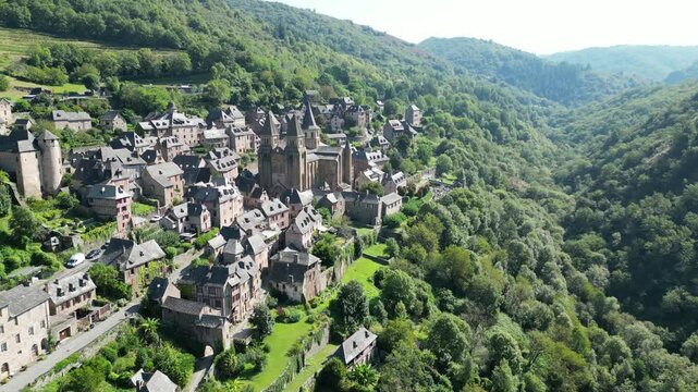 Drone aerial view in France countryside small old medieval town and a cathedral surrounded by a green mountain forest panning in Conques