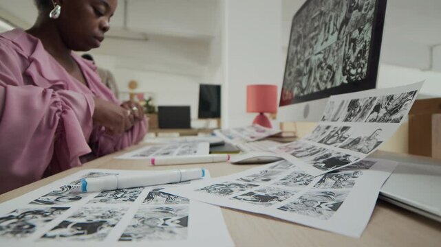 Young Black female graphic designer dressed in pink blouse using colored markers while working on black and white storyboard sheets in front of computer in modern office