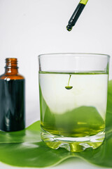 Dropper releasing green liquid into a glass of water with a leaf underneath and a brown glass bottle in the background. Represents natural ingredients, organic products.