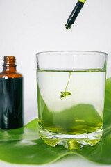 Dropper releasing green liquid into a glass of water with a leaf underneath and a brown glass bottle in the background. Represents natural ingredients, organic products.