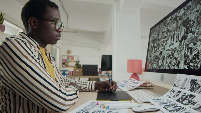 Waist up of young Black male storyboard artist sketching on graphic tablet while looking at computer with draft on screen, sitting at table in modern office setting