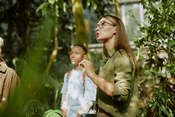 Young female tour guide speaking about rare plants growing in greenhouse while working with group of teenagers