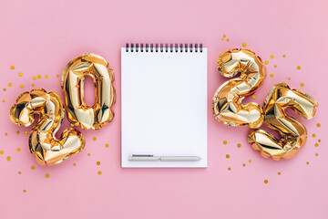 Flat lay of a blank spiral notebook with a pen, surrounded by shiny golden 2025 foil balloons on a...