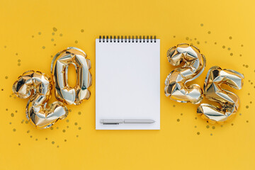 Flat lay of a blank spiral notebook with a pen, surrounded by shiny golden 2025 foil balloons on a...