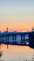 A stunning sunset over the Dnipro River in Kyiv, Ukraine, featuring the Motherland Monument, a bridge, and vivid sky reflections on calm water