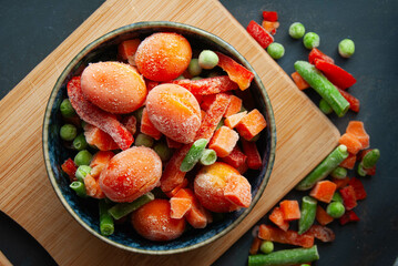 Freshly frozen peas, tomatoes, carrots, and peppers arranged in a bowl, highlighting their textures on a dark blue surface.
