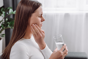 Close up of young woman feel terrible toothache after drink cold water. Sad girl sit on couch in...