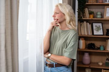 Young sad and depressed woman feel lonely standing in living room looking through the window....
