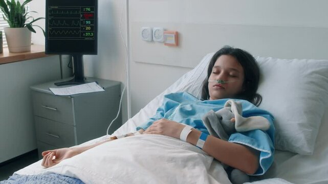 Tilt shot of little Caucasian girl sleeping in hospital bed with bunny plush, with nasal prong and IV in arm during recovery period in clinic