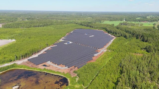 Aerial panoramic tilt down showcasing Solar Farm in Finland with its bifacial panels glistening in sunlight next to open pond in middle of forest