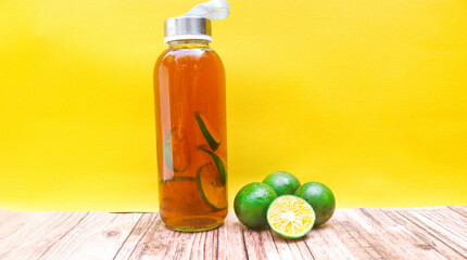Close up bottle of iced tea with lemon, lime, and ice cubes isolated on yellow background and table. A bottle of sweet iced tea full of lemon slices