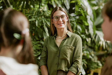 Medium shot of cheerful young female professional botanist showing botanical garden to group of...