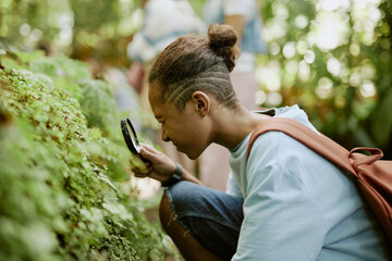 African American teen boy using magnifying glass to look closer at plant growing in botanical...