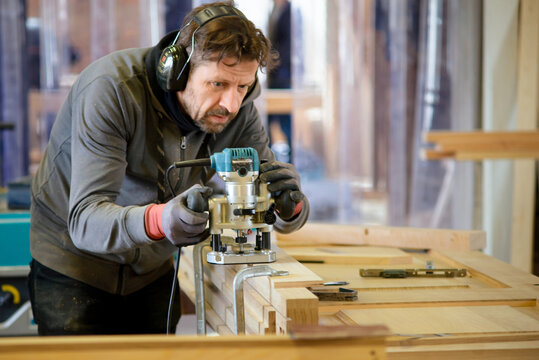 A skilled craftsman works intently in a woodworking shop, using a router to shape and refine wooden materials. Wearing protective headphones and gloves, he focuses on precision and detail.