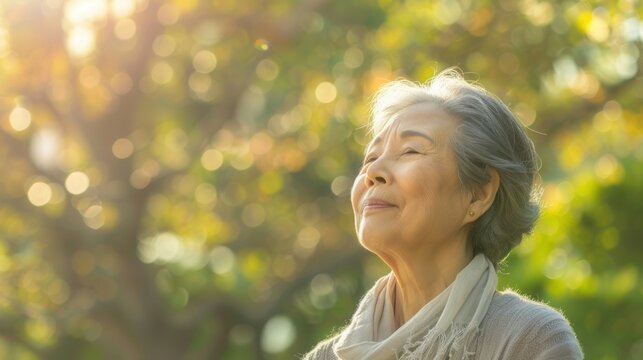 Elderly asian woman with short gray hair smiling while looking up at sunlit tree canopy in natural setting