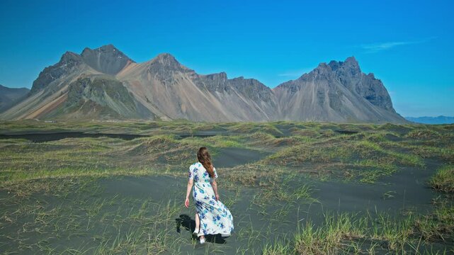 A young woman in a dress enjoys Stokksnes, at Vestrahorn Mountain, Iceland. Tourists visit tall scree mountains made of granophyre rocks. Headland with a dramatic mountainscape and black sands.