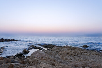 A serene coastal scene at twilight. The rocky shoreline extends into calm waters, reflecting the...