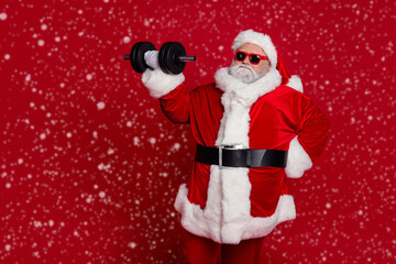 Portrait of attractive bearded funny Santa lifting dumbbell working out difficult hard isolated over bright red color background