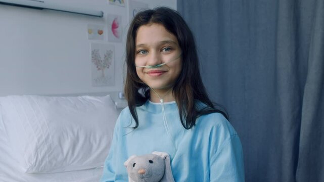 Tilt portrait of little Caucasian girl with nasal cannula hugging grey bunny plush, smiling and looking at camera in ward in childrens hospital