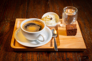 A cup of coffee, a small plate of whipped cream, a glass of water, spoon and a brownie on a wooden tray