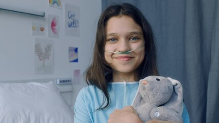Chest up portrait of happy Caucasian 12yo girl with nasal prong embracing grey bunny plush toy in hospital ward decorated with childs paintings and looking at camera