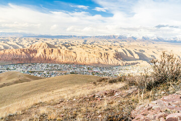 A stunning landscape of a small town at the base of dramatic red cliffs, with layers of rugged mountains in the background under a  blue sky. The scene captures rural serenity and natural beauty