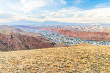 A captivating wide-angle view of a valley surrounded by rugged red rock formations and distant mountains. A town with scattered houses lies below, embraced by nature's dramatic beauty under a sky
