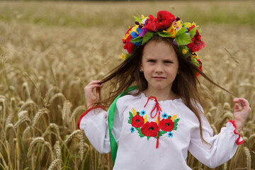 A small sad girl in a colorful wreath and embroidered shirt stands in the middle of a ripe wheat...